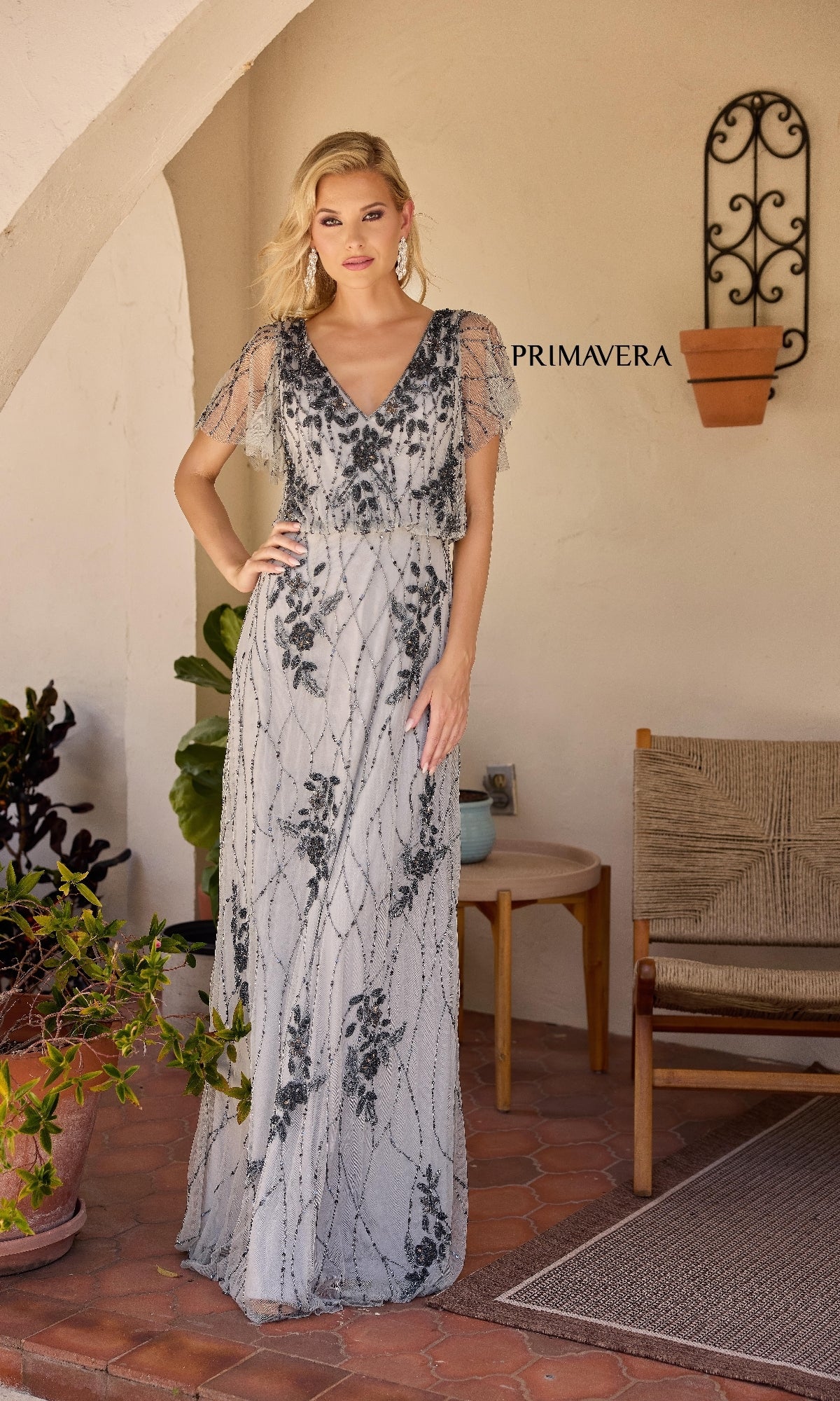 A woman wears the Primavera 13144, a light grey, floor-length formal A-line dress with black floral patterns, standing on a tiled patio near a chair and potted plants beneath an arched wall with a decorative sconce.